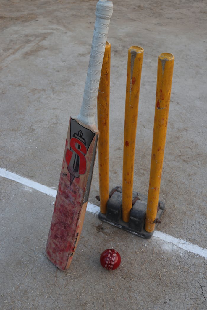 Still life of a cricket bat, red ball, and stumps on a cricket field, highlighting the sport's essential equipment.